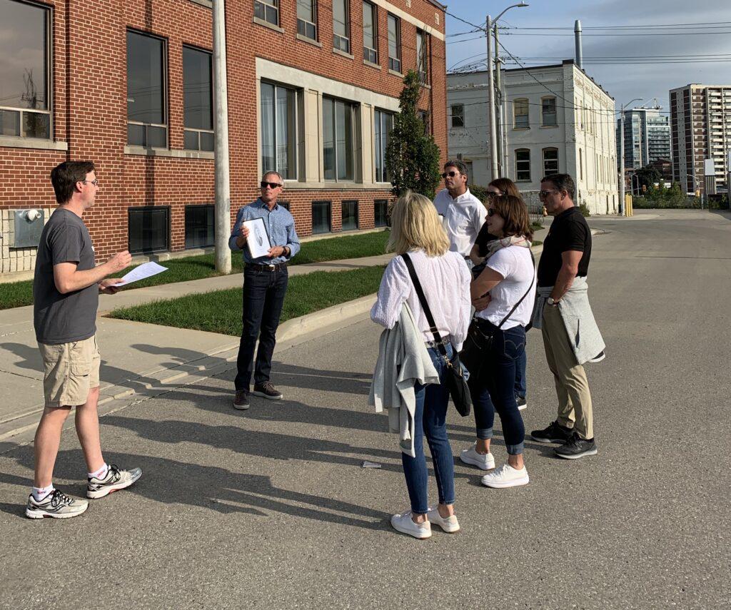 Group of six people on a Stroll Walking Tour facing the guide, Todd Bowman, who is speaking to the group in front of a red brick building in Kitchener's Midtown neighbourhood