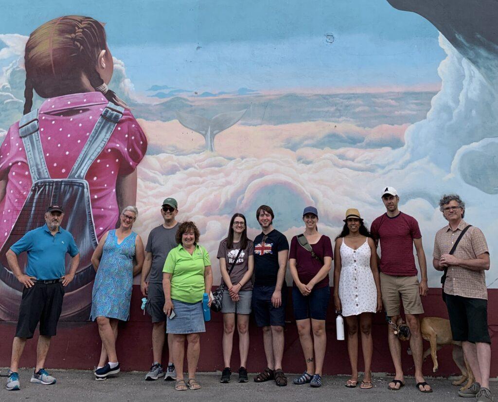 Group of people and one dog standing in front of a mural of two children looking at a whale