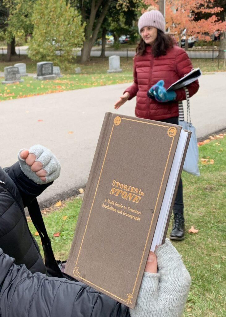 A person's hand holding the book titled Stories in Stone: The Complete Guide to Cemetery Symbolism by Douglass Keister. 