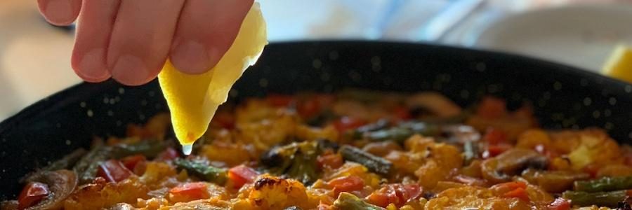 a hand squeezes lemon over paella pan with sun highlighting the lemon